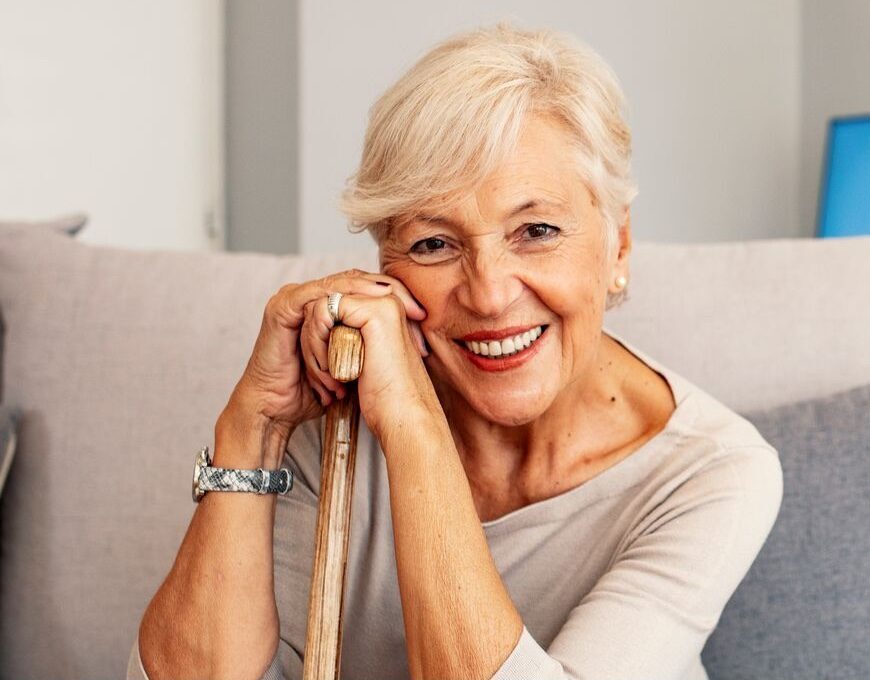 A contented elderly lady sits on a sofa, wearing a warm smile.