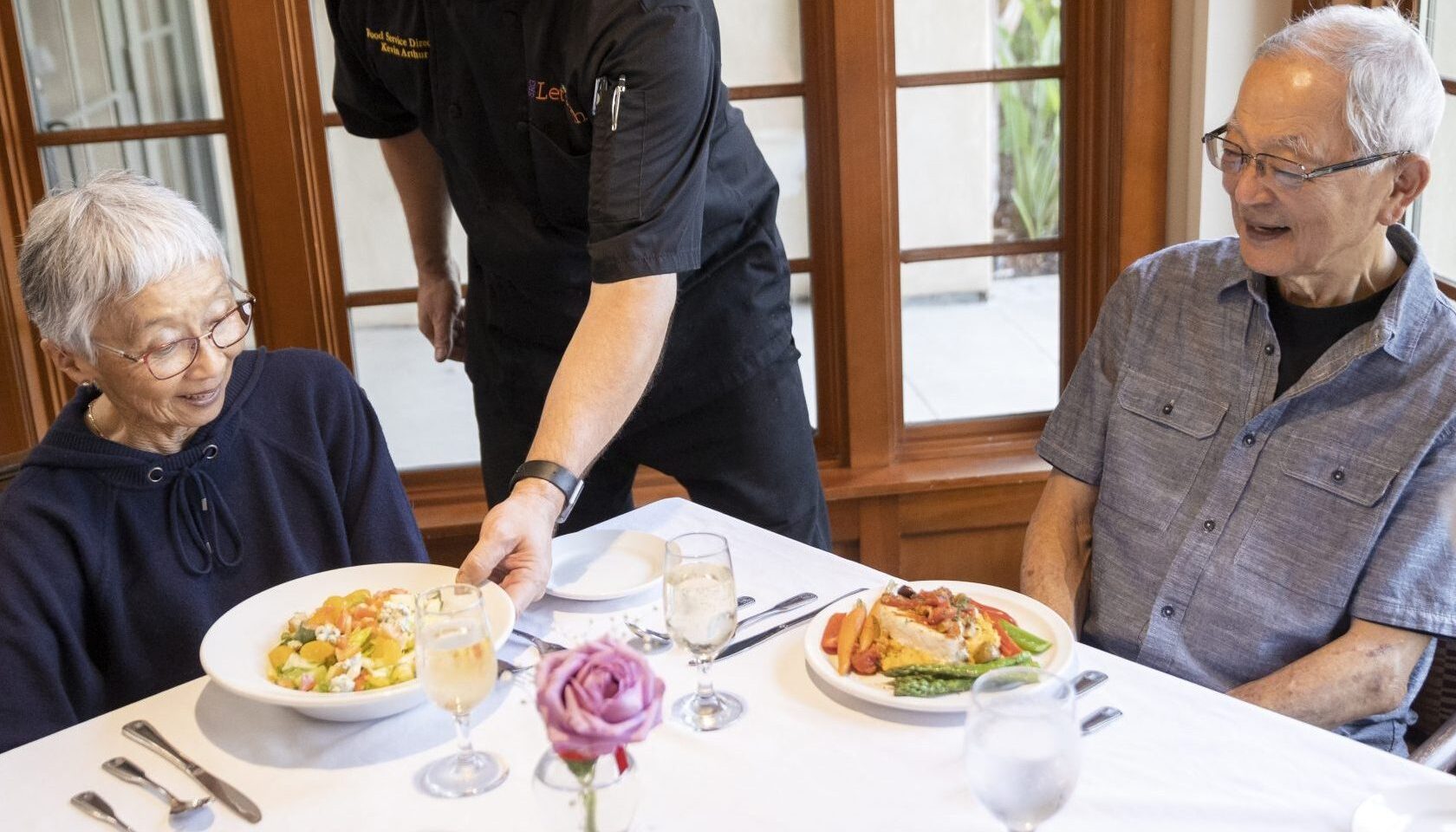 An elderly couple enjoying a meal at a table, with a waiter serving them delicious food.