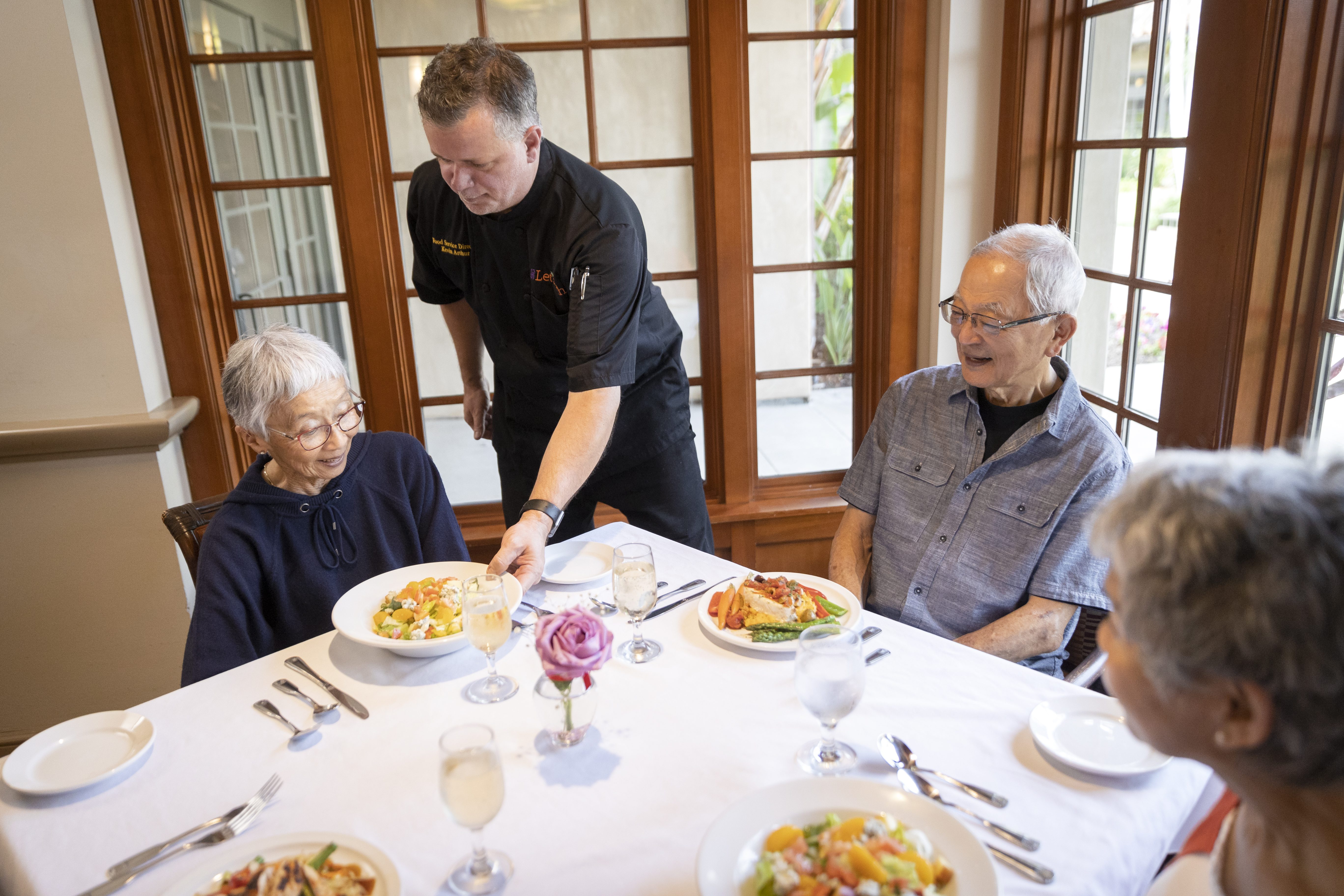 An elderly couple enjoying a meal at a table, with a waiter serving them delicious food.