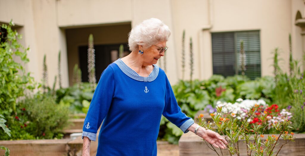 An elderly woman gracefully strolls through a serene garden, wearing a lovely blue dress.