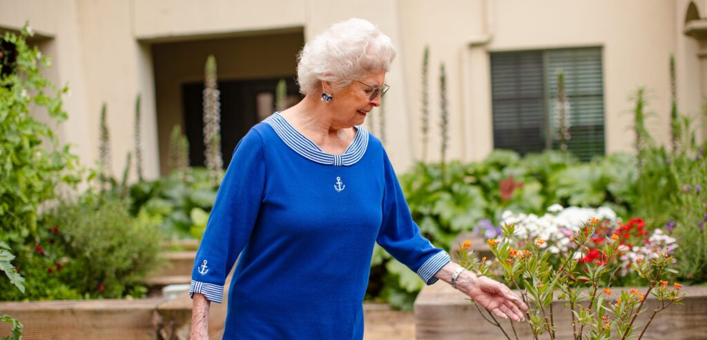 An elderly woman gracefully strolls through a serene garden, wearing a lovely blue dress.