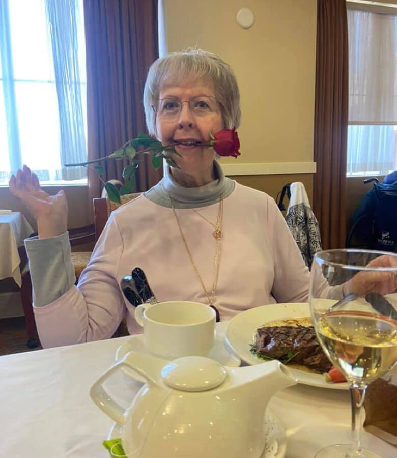 An elegant older woman sitting at a table, gracefully holding a rose in her mouth.