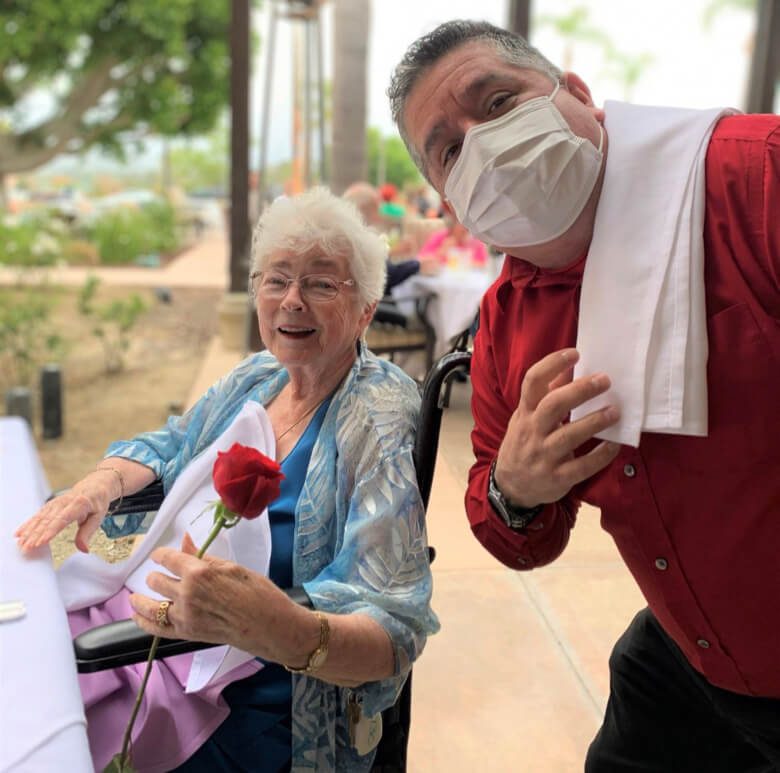 A man wearing a mask stands beside a woman sitting in a chair, holding a rose.