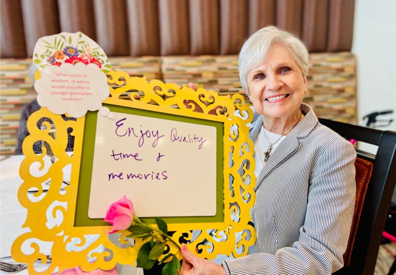 An elderly woman smiling while holding a sign that reads 