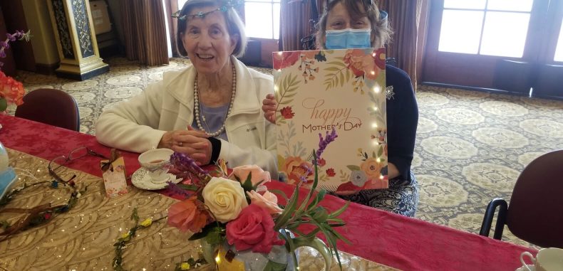 Two women sitting at a table with flowers and a sign, engaged in conversation and enjoying each other's company.