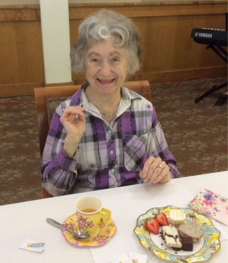 An elderly woman enjoying a meal at a table, with a plate of food in front of her.