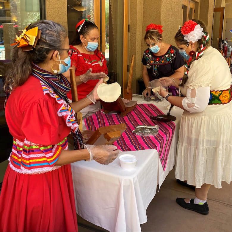 Women in colorful Mexican costumes preparing food at a table for a festive celebration.