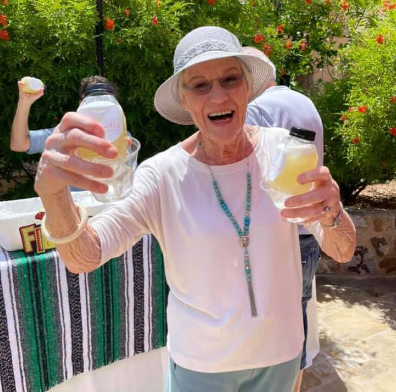 An elderly woman smiling while holding two beverages in her hands.