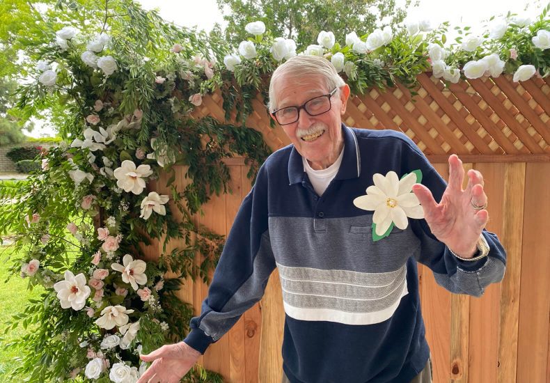 An elderly man gracefully holds a delicate flower, standing in front of a rustic fence.