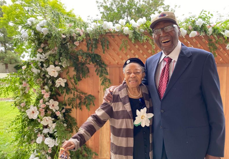 An elderly couple posing in front of a vibrant flower wall, radiating joy and love.