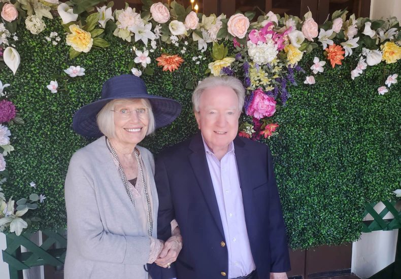 An older couple smiling together in front of a beautiful flower wall.