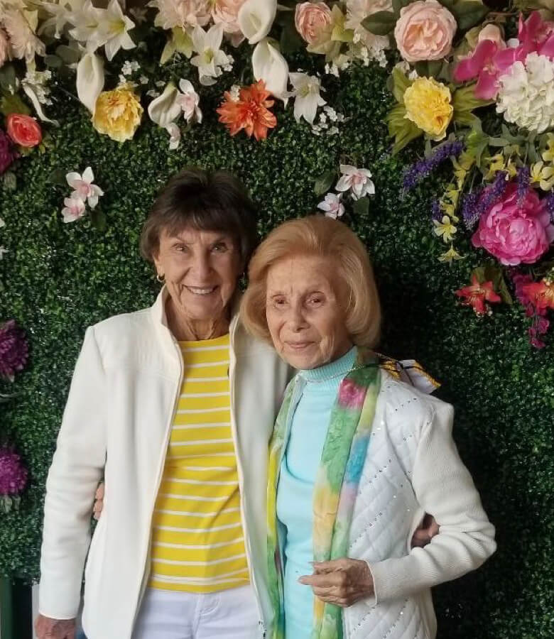 Two women posing in front of a beautiful floral wall, showcasing vibrant colors and natural beauty.