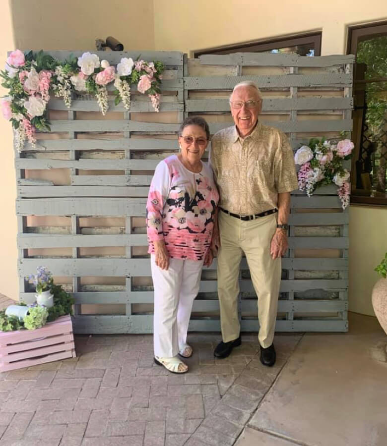 An older couple smiling together against a wooden pallet backdrop, radiating warmth and love.