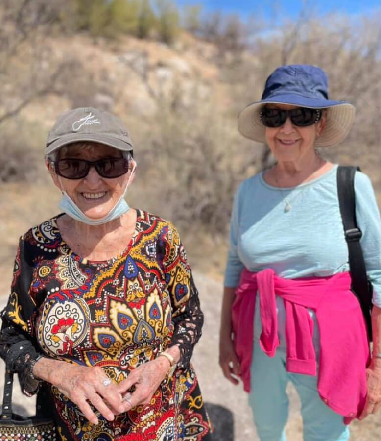 Two elderly women wearing sunglasses and hats, walking on a trail surrounded by nature.