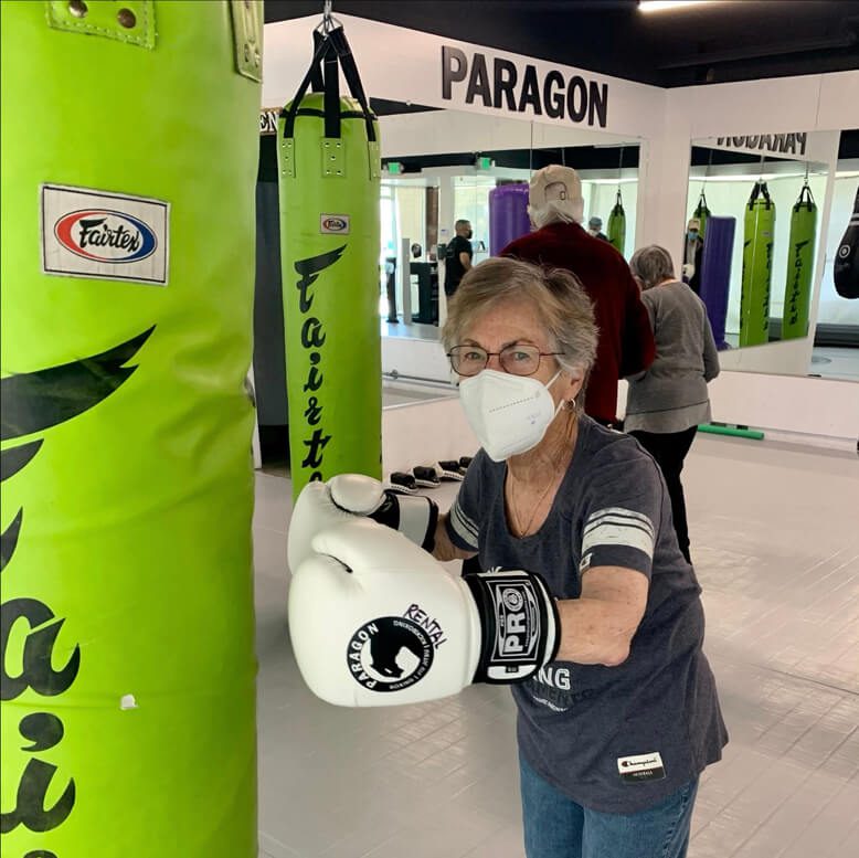 An older woman wearing protective gear in a boxing gym, ready to train with a mask and gloves.