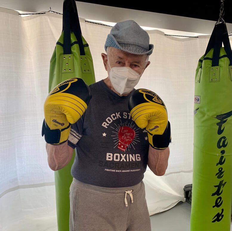 An older man wearing protective gear in a boxing ring, ready to spar with an opponent.