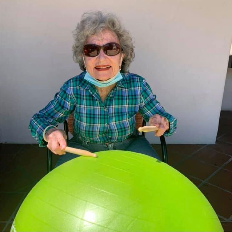 An elderly woman wearing a face mask and sunglasses joyfully playing with a green ball.