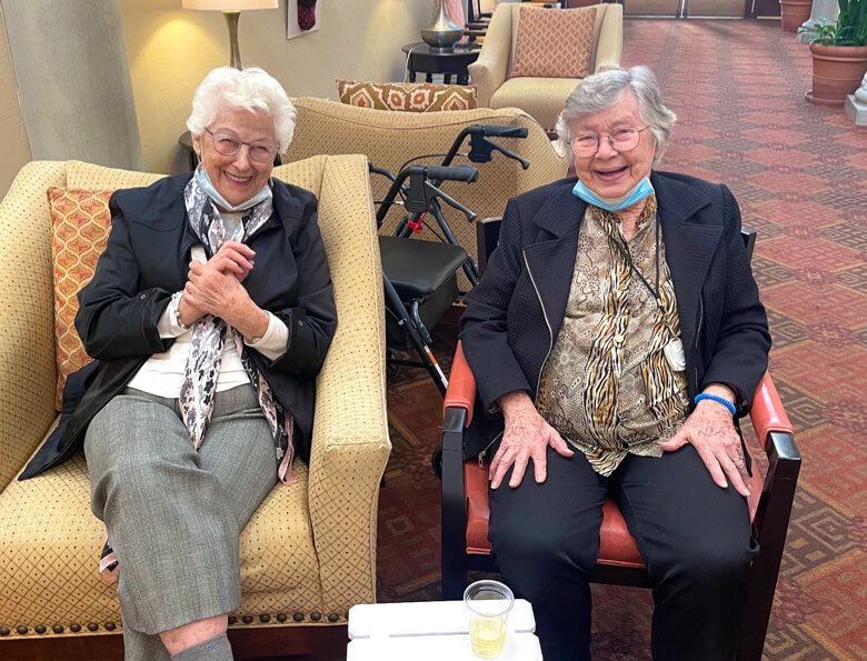 Two elderly women sitting in chairs in a lobby, engaged in conversation and enjoying each other's company.