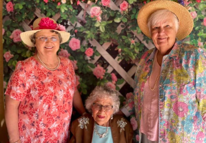 Three women in floral dresses and hats smiling for a photo in a garden.