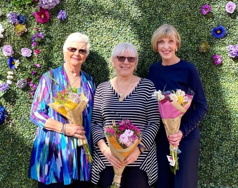 Three women posing in front of a vibrant flower wall, showcasing a colorful and captivating backdrop.