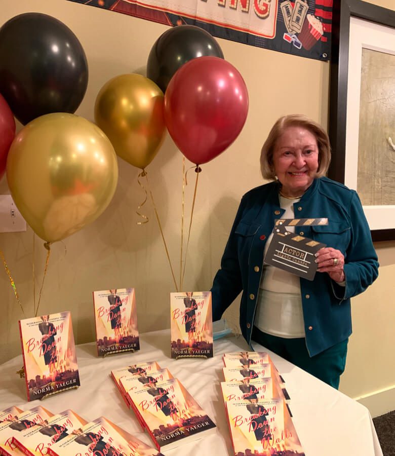 An elderly woman stands beside a table adorned with colorful balloons and books.