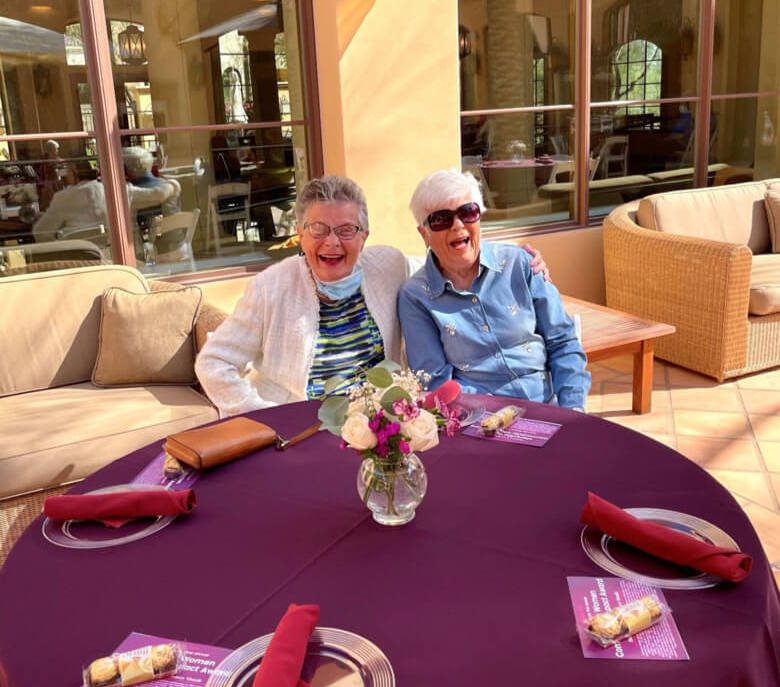 Two elderly women sitting at a table covered in purple tablecloths, engaged in conversation.