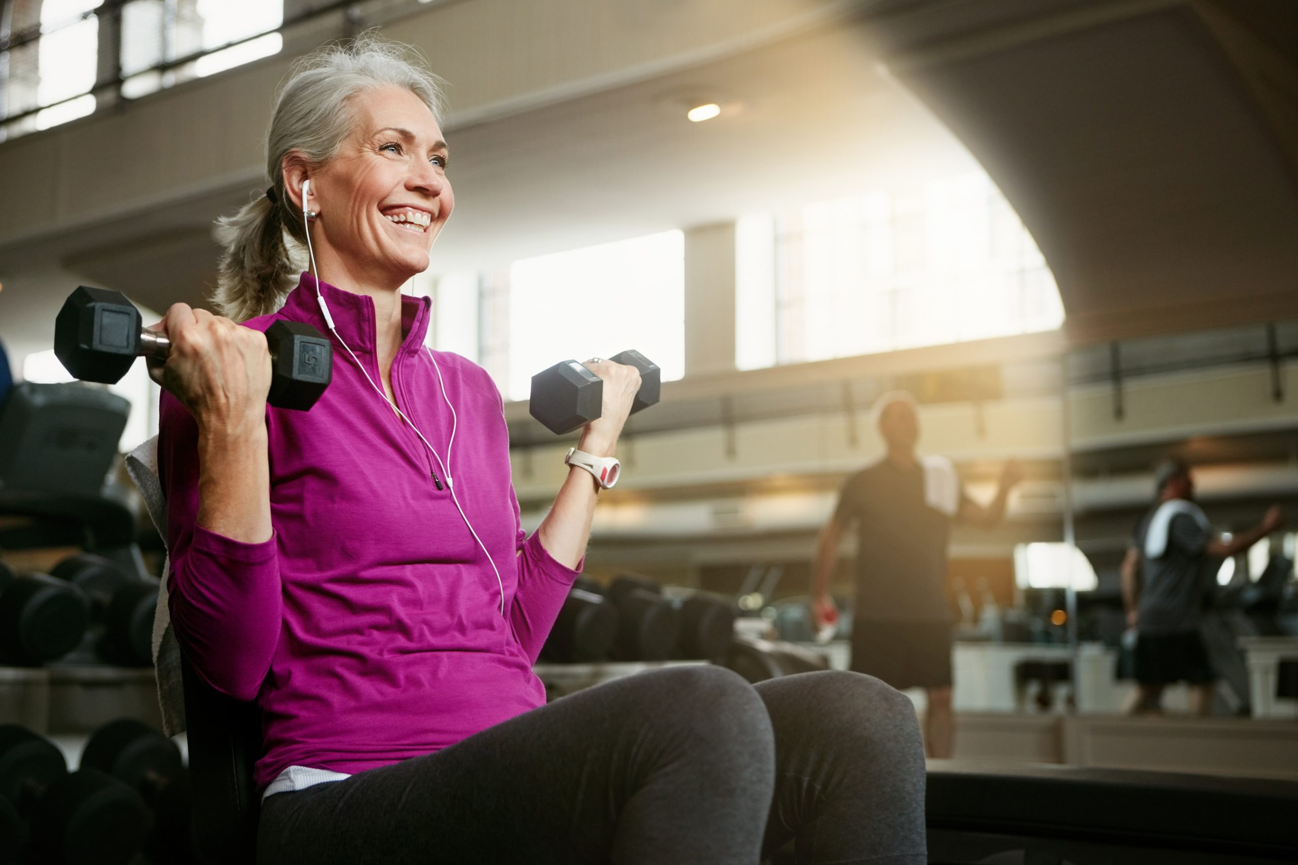 an elderly woman listening to music and doing bicep curls in a gym and smiling