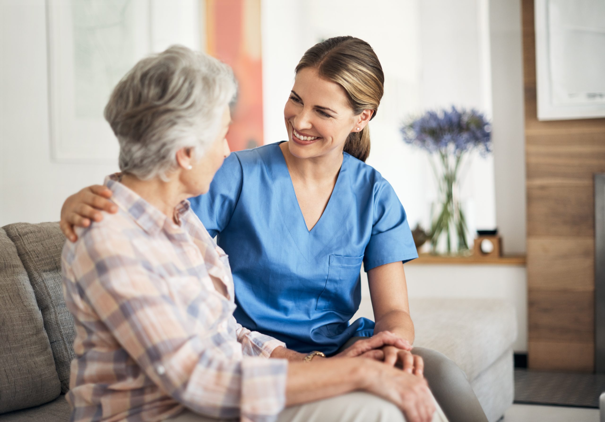 A nurse attentively sits beside an elderly woman on a couch, providing care and support.