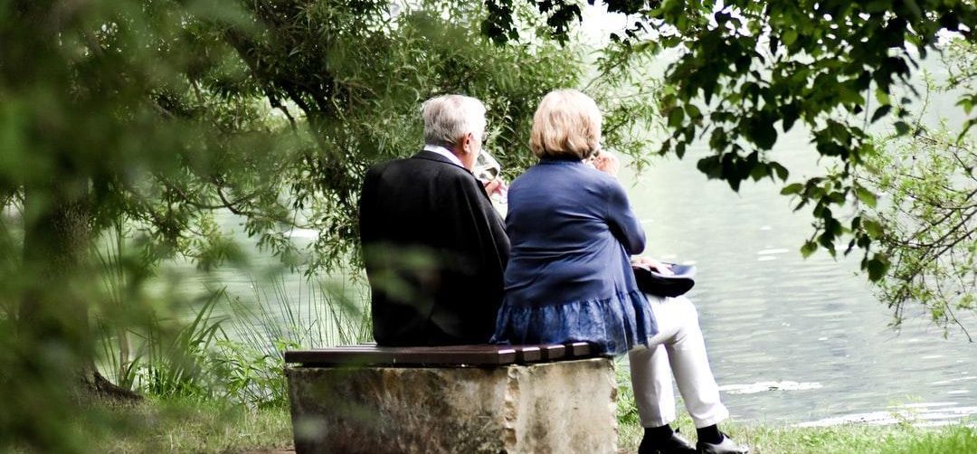 two elderly people drinking wine sitting on a bench next to a river