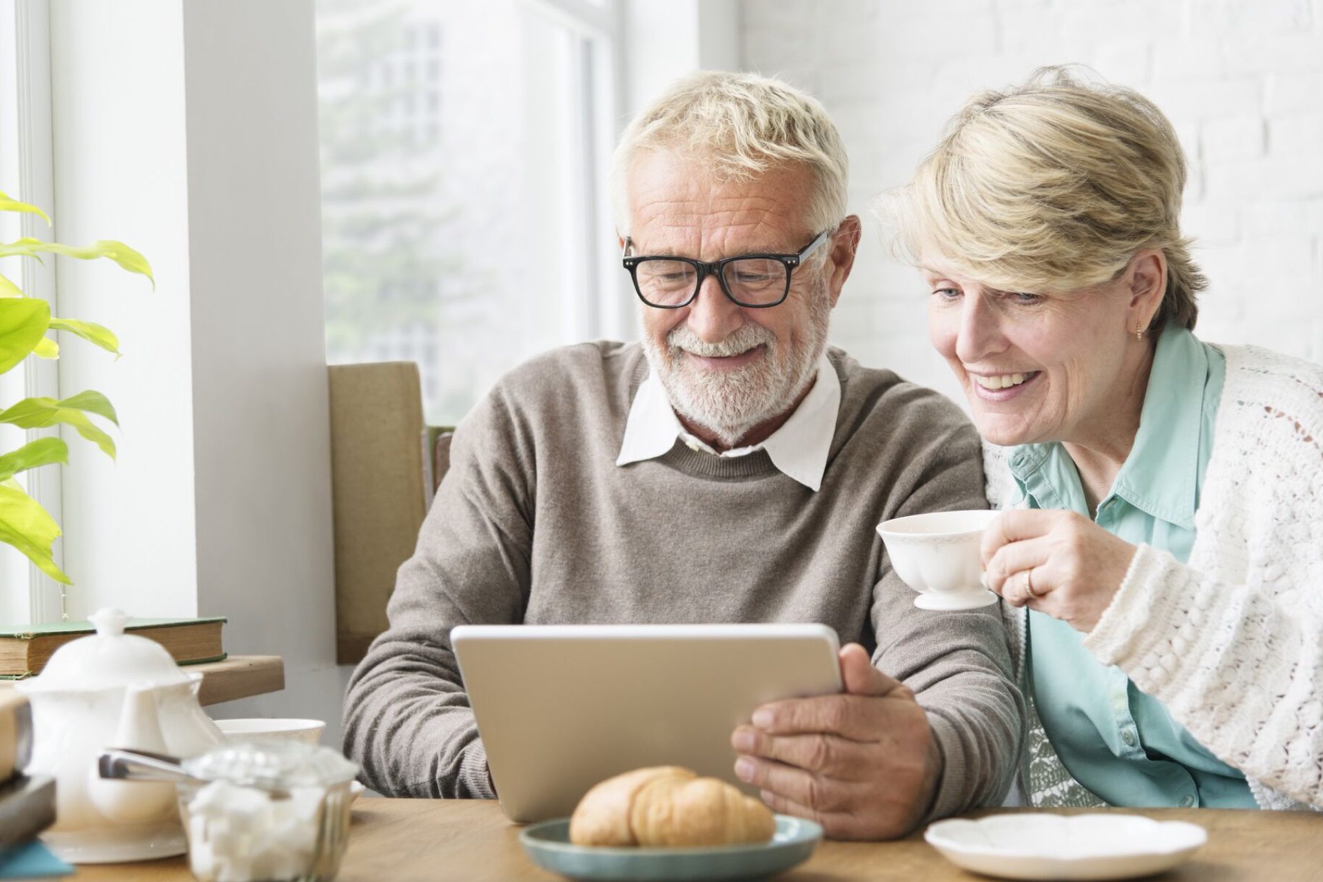 An elderly couple engaged in conversation while sitting at a table, using a tablet device.