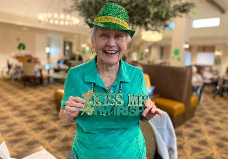 Elderly woman in green shirt and hat holds a sign saying 
